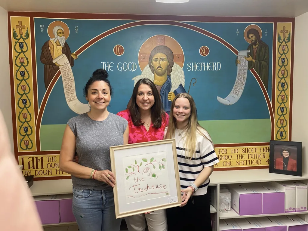 Three women holding a sign saying "The Treehouse" in front of the icon of Christ The Good Shepherd