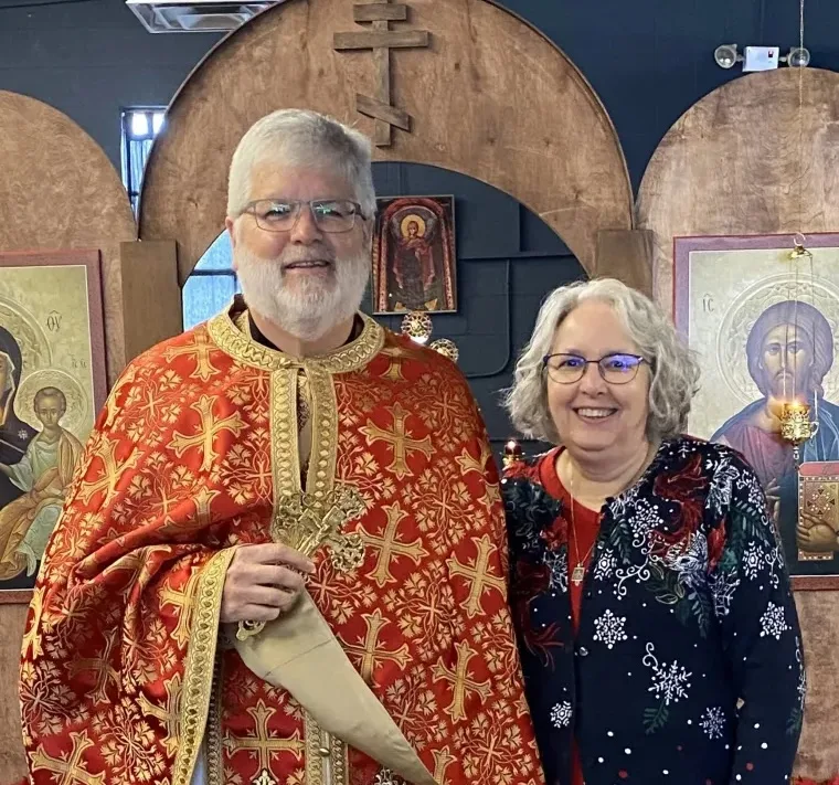 Fr. Andrew dressed in vestments and holding a cross and Kh Belinda in front of the iconostasis